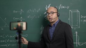 Indian young teacher man wearing headset teaching online video conference live stream by smartphone. Asian teacher teaching mathematics class webinar online for students learning.