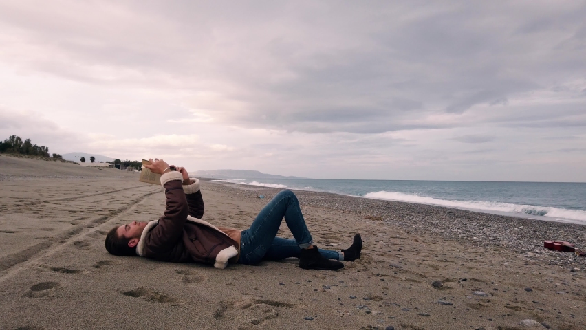 Young boy reads a book sitting on the beach by the sea
