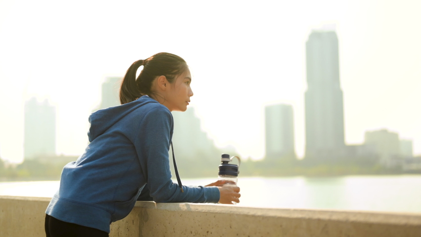 Asian woman drinking water and looking at city view after morning exercise on the bridge.Excercise,running,jogging, fitness and healthy lifestyle concept.  