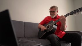 Gray-bearded pensioner with glasses playing guitar at home. Mature man learns to play the guitar online while sitting on the couch near the laptop
