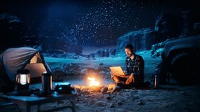 Male Traveler Sitting by Campfire Uses Laptop Computer while Tent Camping in the Canyon. Man doing Digital Remote Work, e-business, ecommerce through Internet while being on Vacation Trip. Static Shot