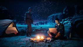 Tent Camping in the Canyon at Night: Man Uses Laptop Computer while Sitting by the Campfire, His Girlfriend Joins Him. They Watch Streaming Service, Share Thoughts, Videos and Photos on Internet