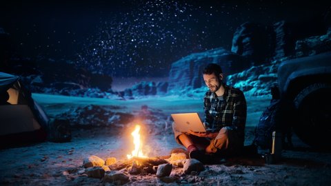 Night Tent Camping in Canyon: Male Traveler Uses Laptop Computer Sitting by Campfire. Man on Digital Remote Work, e-shopping, ecommerce, Using Internet, Social Media Posting on Vacation Trip