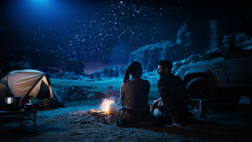 Happy Couple Nature Camping in the Canyon, Sitting by Campfire Watching Night Sky with Milky Way Full of Bright Stars. Two Travelers In Love On a Romantic Vacation Trip. Zoom Out Back View Shot