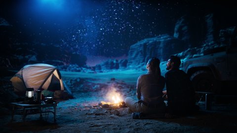 Happy Couple Tent Camping in the Canyon, Sitting by Campfire Watching Night Sky with Milky Way Full of Bright Stars. Two Travelers In Love On a Romantic Vacation Trip. Zoom Out Back View Shot