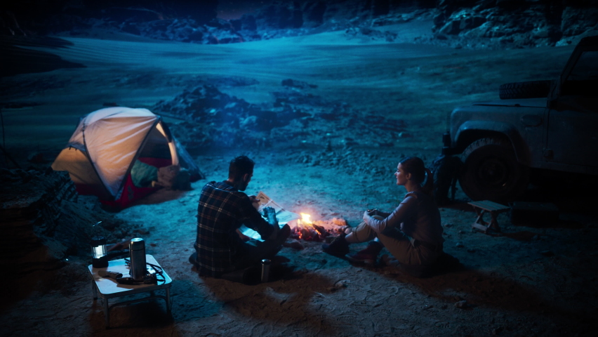Happy Couple of Hikers Camping in the Canyon at Night Together, Sitting by Campfire, Plan Hiking Trekking Journey Route with a Map. Two Traveling people Talk, Milky Way Star Watching. Vacation Trip