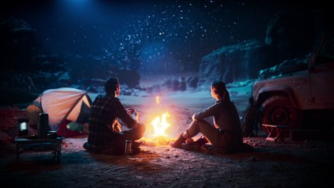 Happy Couple Sitting by Campfire Watching Night Sky while Camping in the Canyon. Two Traveling people Tell Inspirational Stories at Campsite, Look at Milky Way Stars. On Holiday Vacation Trip
