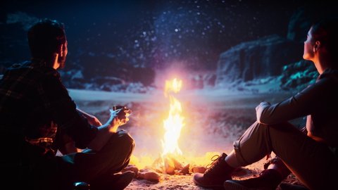 Happy Couple Sitting by Campfire Sharing Cup of Drink, Watching Night Sky while Camping in the Canyon. Two Traveling Young people Tell Inspirational Stories about Life, Look at Milky Way Stars