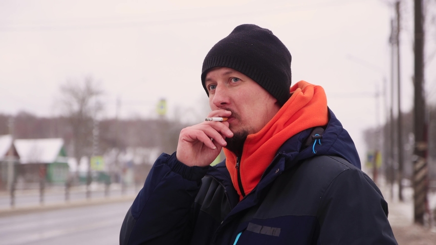 A young man on the side of the road smokes and looks at the passing vehicles. A male in a warm jacket with an orange collar stands on the highway. Cars are passing in the background in defocusing. | Shutterstock HD Video #1067851154