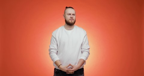 Bearded young man in white sweater, jeans with hair in tail isolated on red background. People emotions lifestyle concept. He is looking at camera and showing thumb down dislike gesture and saying no.