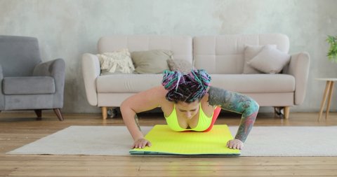 Plump woman newcomer in sport wearing in bright sportswear is doing push-ups exercise in living room standing on knees on mat. Joke, mem, humor, parody. Home workout, training, fitness concept.