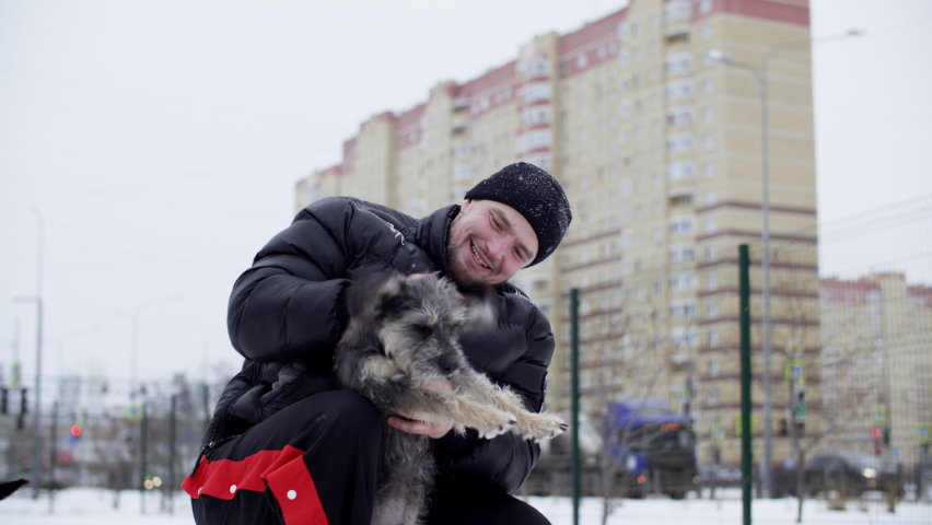 Happy man stroking fluffy dog on winter walk on snowy city landscape. Cheerful dog owner scratching happy puppy on snowy street