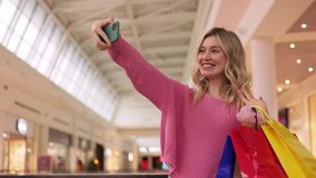 Blonde girl making selfie by smartphone while holding colored gift bags. Blogger influencer speaking to camera phone recording vlog for channel.Smiling female talking in video conference by smartphone