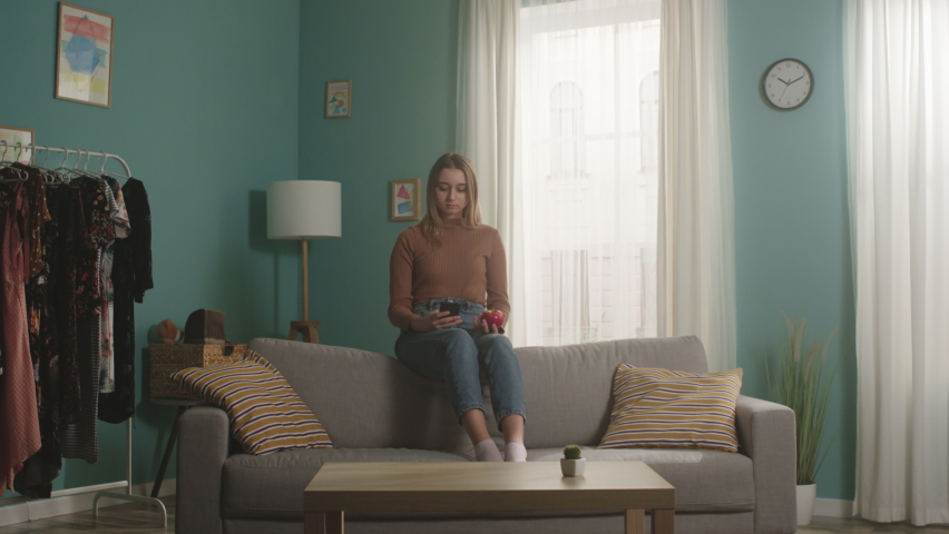A young woman in blue jeans and a brown sweater sits on the back of a sofa, holds a red apple and takes a selfie