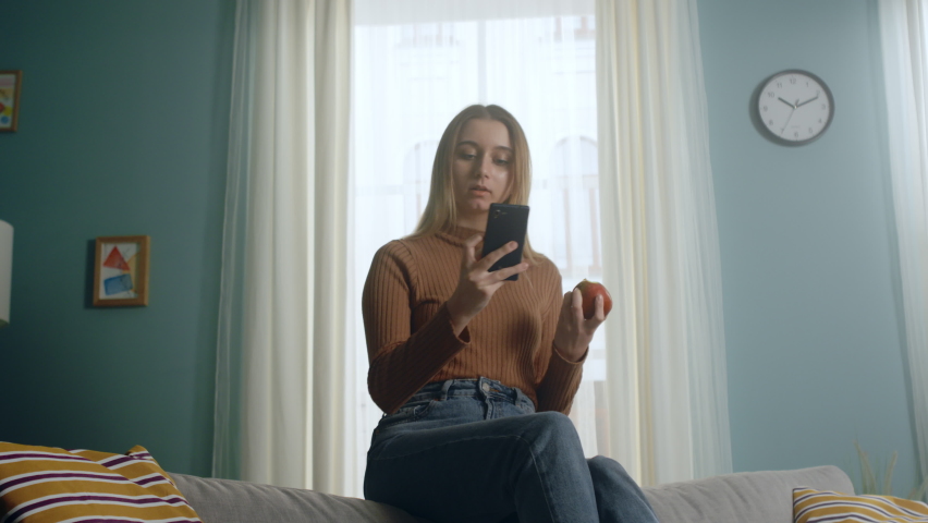 Young woman in blue jeans and brown sweater sits on back of sofa on background of window, holding red apple and taking selfie. Clock hangs on wall. Average plan.