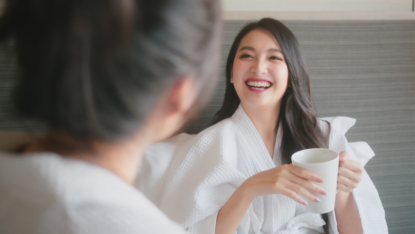 Casual relax holiday morning asian female friends in white bathing suit hand hold hot coffee drink laydown on pillow bed happiness conversation talk on bed in hotel ,casual girlfriend talk | Shutterstock HD Video #1067871686