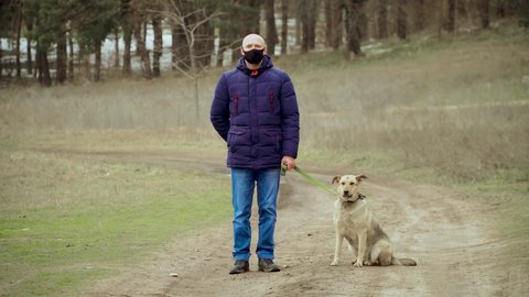Male in black protective anti corona virus face mask motionless stand outdoors with dog on leash in near forest in winter during self isolation on quarantine. Stop dangerous COVID-19 infection
