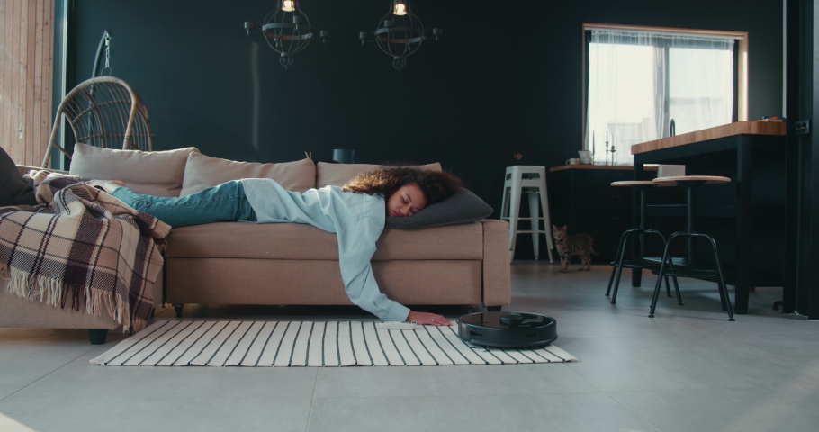 Young tired mixed race freelancer woman sleeping on couch at home, robot cleaner sweeping floor, cat watching scared. | Shutterstock HD Video #1067875409