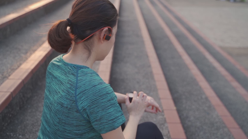 asian athlete woman wearing headphone using sport smart watch sitting on stair at sport club stadium in evening sunset. female in sportswear touching screen application tracking activity and music. 
