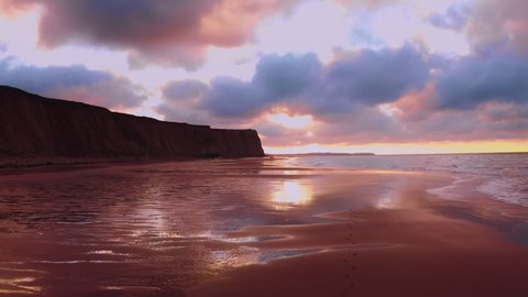Aerial view drone next to a cliff on a golden sunset beach with sun rays peering through the purple blue clouds creating reflections over a dark foamy seawater: film stockowy