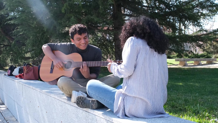 Young latin man playing the guitar and his friend recording him with the smartphone at the university campus. University life, millennial generation.