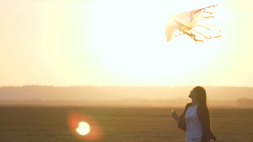 Girl plays with a multi-colored kite on field in rays of sun. Teenager dreams of flying and becoming pilot. colored kite hangs in air. Kite Festival. Child and flying kite at sunset in summer.