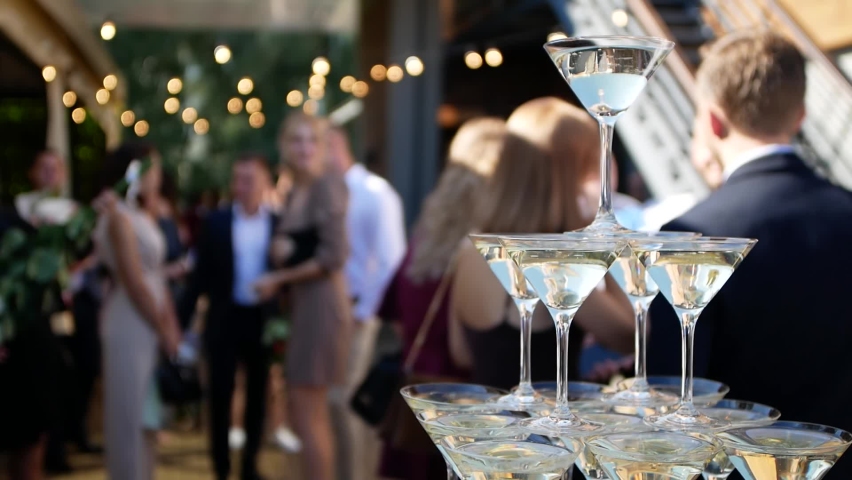 Close-up shooting pyramid of glasses of champagne stands on the buffet table against the backdrop of a cheerful crowd of guests. | Shutterstock HD Video #1067882834