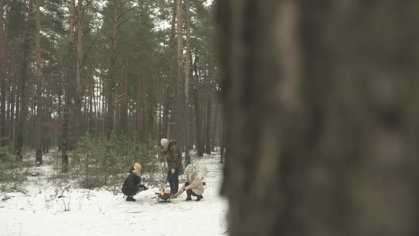 A company of young people are resting in the winter forest and preparing bread and sausages on the fire