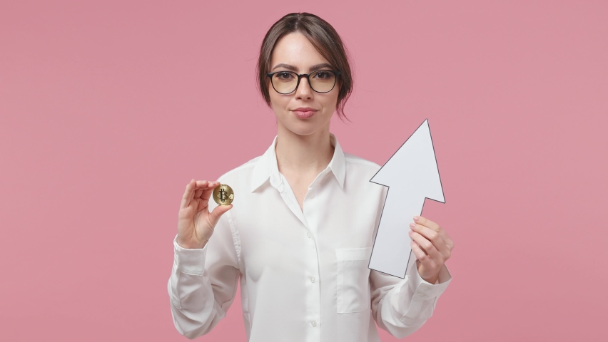 Excited joyful young brunette business woman 20s years old in white shirt glasses posing isolated on pink background in studio. Achievement career wealth concept. Hold up arrow bitcoin coin currency