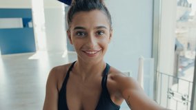 Young beautiful female ballerina standing in gym and talking on video call with friends. View from camera.