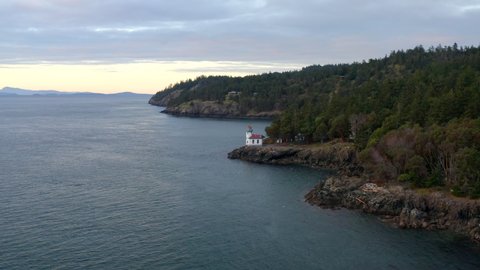 Drone flying towards lighthouse at sunset in the Pacific North West San Juan Islands. Birds flying over ocean with forest in background.