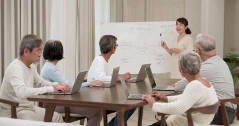 A beautiful kindness young Asian nurse standing near white board and teaching life skills of older to senior male and female and they try to take notes with their computer in classroom at nursing home