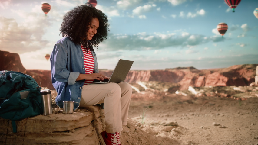 Excited Beautiful Young Multiethnic Female Traveller with Afro Hairstyle Using Laptop Computer on Top of Rocky Canyon Valley. Hot Air Balloons Fly in Mountain National Park. Hiking in Nature.