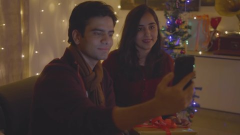 close up shot of a young attractive Indian cheerful couple sitting at home on a couch together are smiling and interacting with a friend or family using mobile phone online video call in a house