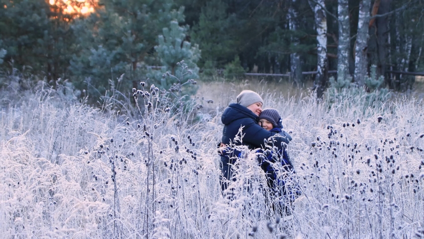 Happy boy and mom run towards each other and happily hug. Family in the winter outdoors. People walk through the snow-covered forest in the evening at sunset. Beautiful fabulous natural landscape.