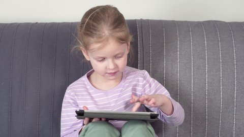 Curious cute little girl using digital tablet to learn while sitting on the sofa in the living room at home.