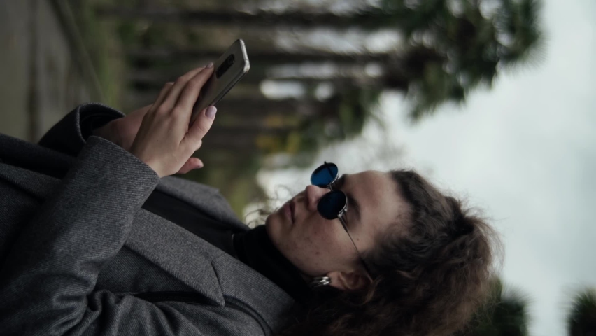 Young stylishly dressed woman in blue glasses looks into the phone. Vertical shot of a girl standing on the street under palm trees. Cool colors. Slow motion 