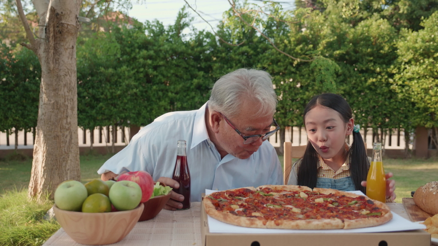 Asian retirement grandfather and pretty granddaughter enjoying to eating pizza together in home garden. Happy senior life after retirement with family concept, S3niorLife