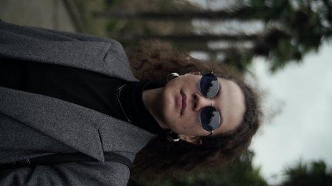 A curly young woman with long curly hair gathered on her head looks into the camera. Cool stylish urban outfit with sunglasses and a gray coat. Neck jewelry Adlı Stok Video