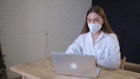 A remote employee wearing a medical mask holds a conference call with a colleague in a virtual online chat using an application launched from the home office. Classic office wear.