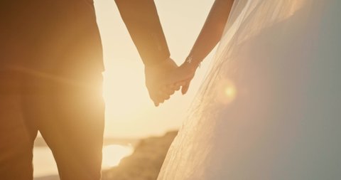 Wedding couple holding hands on sunset background. Close up. Beautiful bride and groom near the ocean.Honeymoon romantic couple in love walking in nature. Lovers or newlywed married by the sea.
