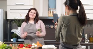 Attractive plus size woman dancing and preparing vegetable salad while her friend is filming video for social networks on smartphone in the kitchen. Blogging and living online concept.