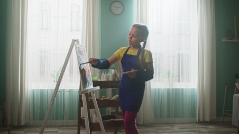 Schoolgirl in blue apron is practicing painting at home, making several brush strokes, than is looking at camera and smiles, Slow motion.