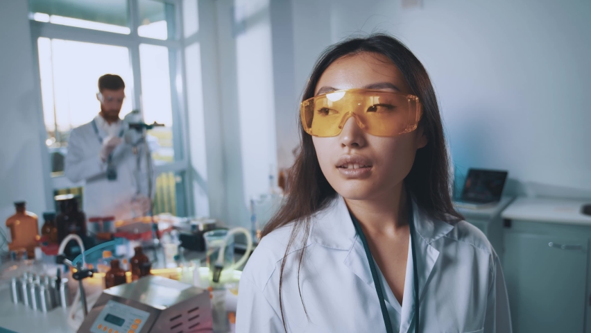 Beautiful young asian scientist woman look at camera in protective glasses. Background team work in the laboratory. Pharmacy, microbiology, industry, biology, technology, doctor. Slow motion | Shutterstock HD Video #1067936618