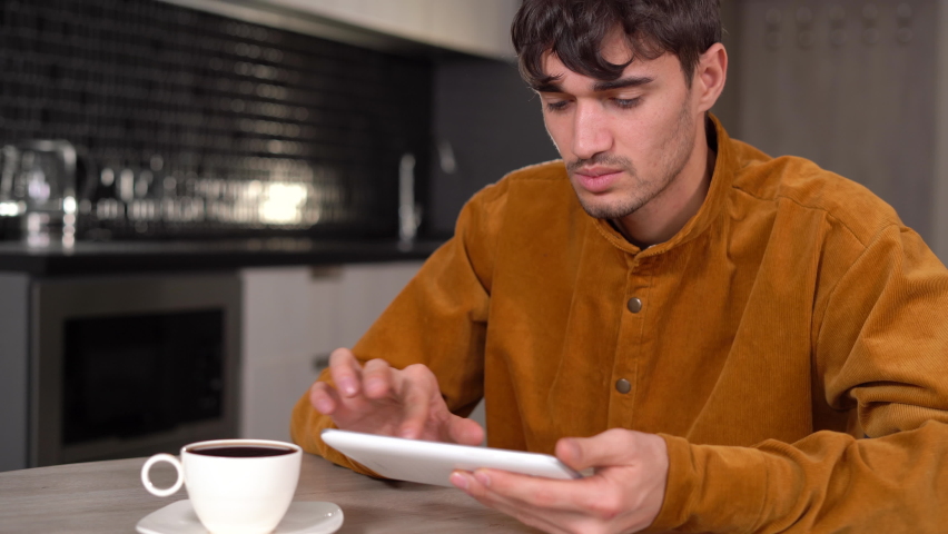 A man in the kitchen, having breakfast and drinking tea or coffee from a cup, sends a message from a tablet and smiles. social network