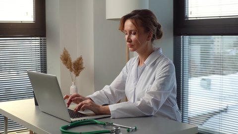 Focused young female doctor in white coat typing on laptop computer looking on display screen, sitting at desk in office room at hospital near window. Woman physician working online and taking notes.