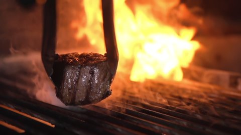 Cooked steak mignon is taken from a hot, burning grill. Metal tongs on a macro plan in slow motion