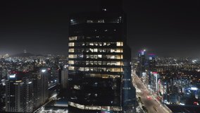 Close up aerial view to a skyscraper in Seoul Gangnam District at the night. Camera moving up high around the building showing illuminated offices inside and beautiful city panorama on the background.