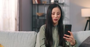 Close up portrait of Asian young beautiful happy woman sitting on couch at home in room speaking on video call online on smartphone. Pretty joyful female videochatting on cellphone, talk concept