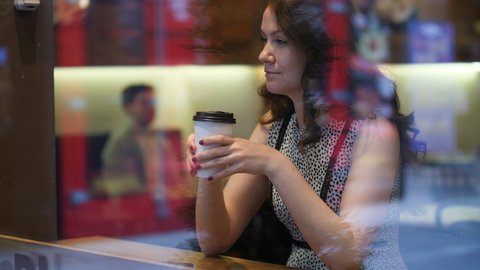 Dreamy woman sitting in cafeteria, look out from window. Shoot through glass with reflection, blurred background. Pretty lady stare at distance, hold cup in hands
